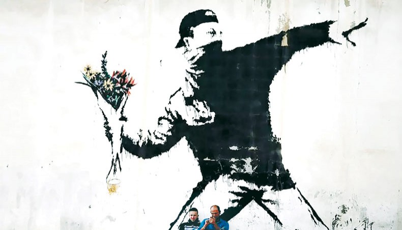 Two men sit in front of Banksy’s ‘Flower Thrower’ mural painted on a wall of a petrol station in the West Bank town of Bethlehem. — AFP photo